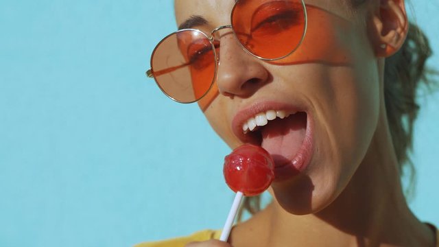 close-up of face of a young tanned laughing woman. cheerful smiling woman with red eyewear standing against blue background and joyfully licking a big red lollipop. Female bright make-up for lips