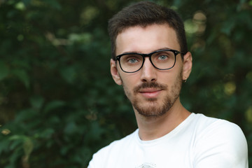 Portrait of a young man in glasses and a white t-shirt on a background of foliage.