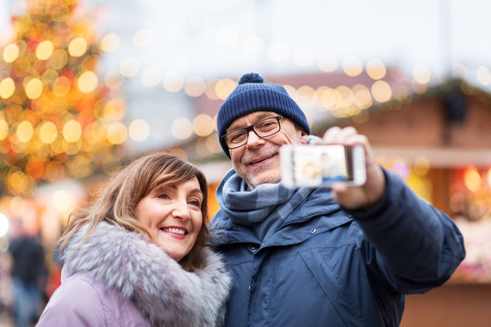 technology, winter holidays and people concept - happy senior couple taking selfie by smartphone at christmas market on town hall square in tallinn, estonia
