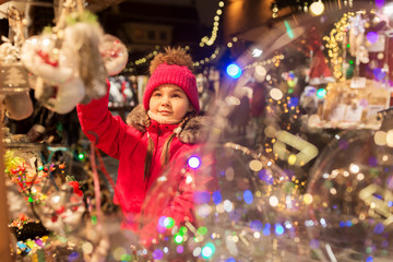 Fototapeta na wymiar holidays, childhood and people concept - happy little girl choosing christmas decorations at market in winter