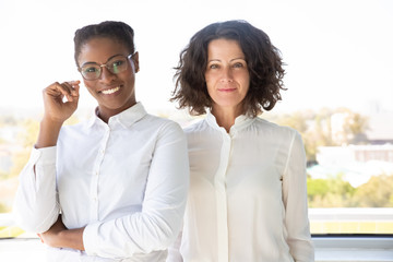 Happy female business partners posing together near office window. Two diverse businesswomen standing for camera and smiling. Teamwork concept