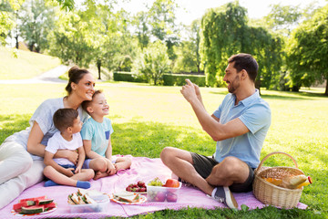 family, leisure and people concept - father taking picture of happy mother with two little sons by smartphone on picnic at summer park