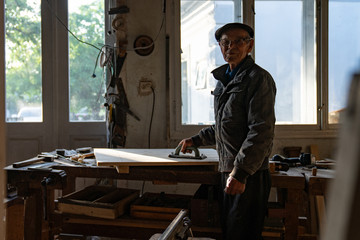 Portrait of very old carpenter in glasses in grey clothes and hat that posing near the workplace holding grinding tool in hand