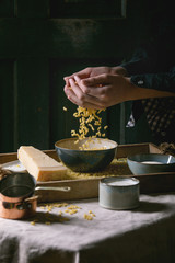 Young man in black sprinkling raw uncooked italian pasta coquillettes for cooking dinner with parmesan cheese, cream and semolina flour on wooden tray on kitchen linen table cloth. Dark rustic style.