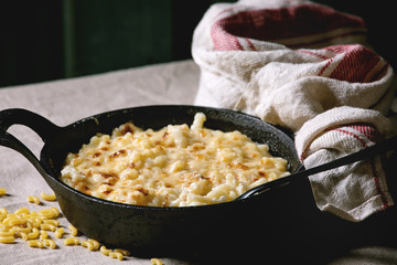 Classic american dish baked mac and cheese in cast iron pan with kitchen towel and ingredients above on kitchen table with linen tablecloth. Dark rustic style