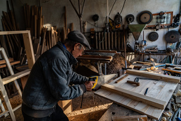 Very old master carpenter in glasses and grey clothes working hard in his home workshop, sawing board