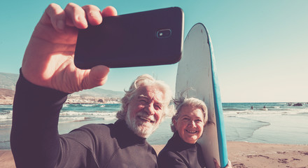 happy couple of seniors at the beach trying to go surf and having fun together - mature woman and man married taking a selfie with the wetsuits and surftables with sea or ocean at the background