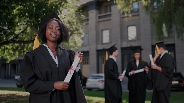 Portrait of the African American cheerful young graduated woman posing to the camera and showing diploma in front of the University. Celebration of pass exams and get trophy status diploma.