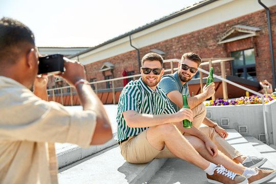 leisure, male friendship and people concept - man with camera photographing his friends drinking beer on street in summer
