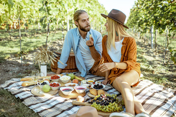 Beautiful couple having romantic breakfast with lots of tasty food and wine, sitting together on the picnic blanket at the vineyard on a sunny morning