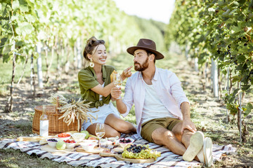 Beautiful couple having romantic breakfast with lots of tasty food and wine, sitting together on the picnic blanket at the vineyard on a sunny morning
