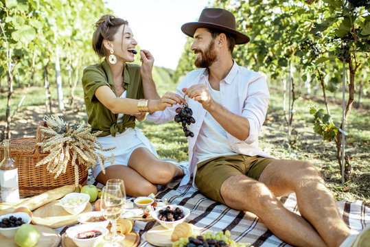 Beautiful couple having romantic breakfast with lots of tasty food and wine, sitting together on the picnic blanket at the vineyard on a sunny morning