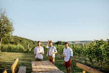 Three guys dressed casually tasting wine while spending time together on the vineyard on a sunny summer morning, landscape view with copy space