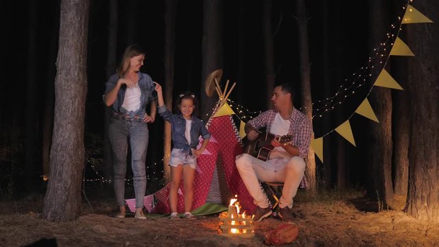 carefree childhood, happy beautiful girl dancing with her loving mother to music of her father playing guitar during night picnic in forest backdrop of wigwam