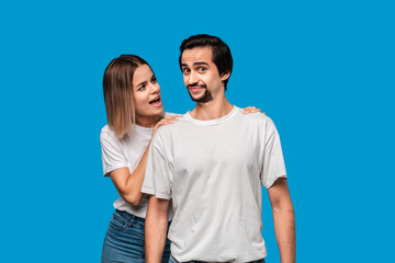 Fototapeta premium Young blond woman tells somthing important to her brunet bearded boyfriend with mustaches in white t-shirts and blue jeans isolated over blue background. Concept of perfect couple.