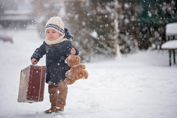 Baby playing with teddy in the snow, winter time. Little toddler boy in blue coat, holding suitcase and teddy bear, playing outdoors in winter park