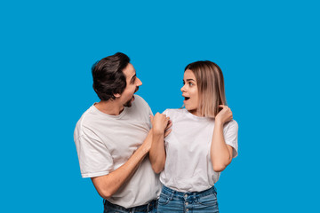 Fototapeta premium Happy couple of a young blond woman and brunet bearded man with mustaches in white t-shirts and blue jeans posing isolated over blue background. Ideal couple.