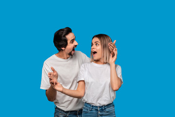 Happy couple of a young blond woman and brunet bearded man with mustaches in white t-shirts and blue jeans posing isolated over blue background. Ideal couple.