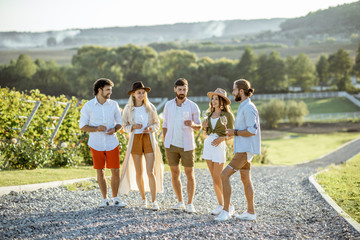 Group of young friends dressed casually hanging out together, walking with wine glasses on the vineyard on a sunny day