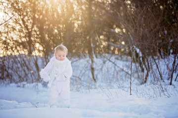 Baby playing with teddy in the snow, winter time. Little toddler boy in handmade white snowsuit, holding teddy bear on sunset, playing outdoors in winter park