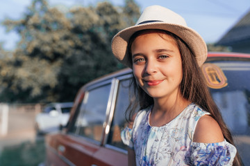 Clsose up portrait, beautiful young brunette little girl in dress and hat leaned on the red old antique car and looking at the caamera