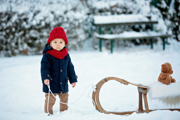 Baby playing with teddy in the snow, winter time. Little toddler boy in blue coat, sliding on...