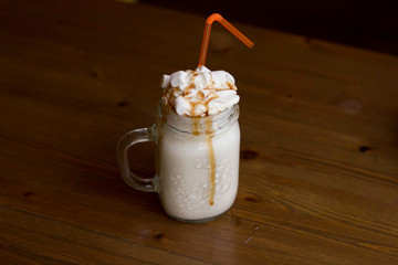 Milk  shake in glass jar with whipped cream, marshmallow, pumpkin and caramel syrup, orange straw on wooden table, autumn fall October mood, with yellow pillow on background. Dark food beverage photo