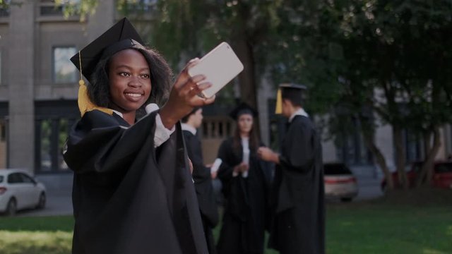 Young african american female graduate making selfies on smartphone camera. Pretty and clever girl with diploma in academic cap and mantle or gown.
