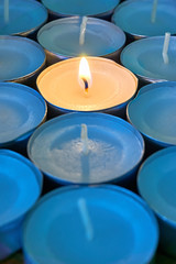 Closeup of tea lights with one being lit. A single warm orange flame between  candles in blue shadow. Christmas, mourning, condolence, memorial, funeral or cremation ceremony concept. Vertical image  
