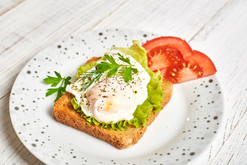 Poached egg, benedict on toasted white bread toast with salad and spices on a plate on a light wooden background.