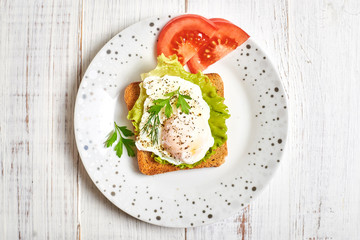 Poached egg, benedict on toasted white bread toast with salad and spices on a plate on a light wooden background.