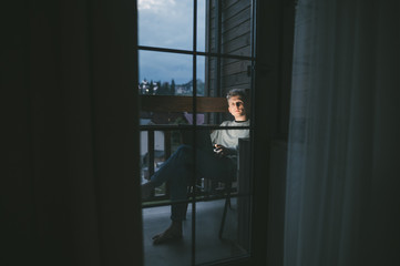 A young man in the evening sits on a balcony in an apartment with a laptop on his lap and looks away. Evening portrait of a guy working on a laptop on the balcony in the evening. Freelance