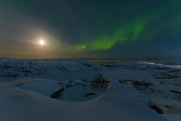 Northern lights, aurora in the sky at night. Hills and rocks covered with snow. Moon in the sky.
