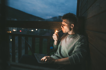 Handsome young man sits at night on the balcony with a laptop on his lap and lights a cigarette with a lighter.Evening portrait freelancer smokes and works on a laptop on a balcony in an apartment