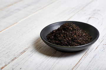 Black dry tea in a clay bowl on a light wooden background.