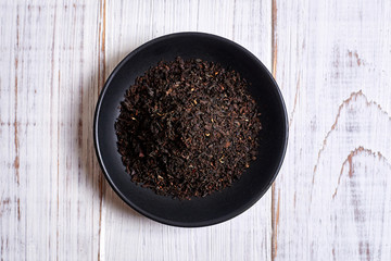 Black dry tea in a clay bowl on a light wooden background.
