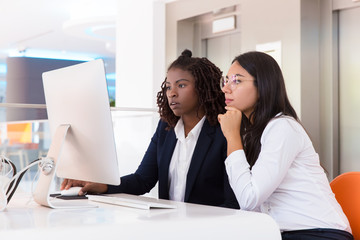 Female office colleagues watching project presentation together. Young business women sitting at table, using computer, looking at monitor. Computer technology concept