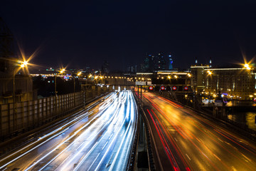 Fototapeta premium Panoramiczny widok na miejską autostradę ze świecącymi liniami światła reflektorów i architekturą na tle nocnego ciemnego nieba