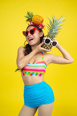 Beautiful woman in a swimsuit holding a pineapple poses on a yellow background.