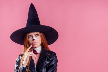 redhaired ginger woman witch black hat and in leather jacket take thought and scaring with crazy make up on her pretty face in studio pink background