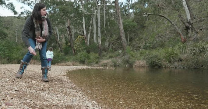 woman on a stony riverbank attempting to skip stones across the creek while her children play in the background. woman throwing stones into a river, beautiful tranquil bush creek
