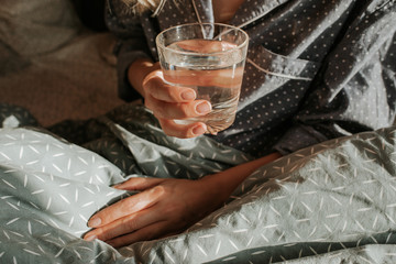 Woman in bed holding a glass of water in hand. Happy morning. Girl in pajamas. Healthy lifestyle, wellness. Proper nutrition. Drinking water. Morning with water. Sunlight on linens. Pillow, blanket