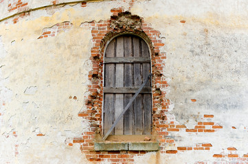 Fototapeta na wymiar window of an old abandoned church boarded up with wooden boards and a wall with crumbling plaster