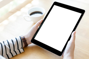 Hands holding black digital tablet pc with white blank screen on wooden table background in cafe.