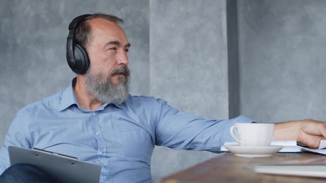 Tracking left of grey-haired bearded businessman sitting at desk, analyzing documents and making amendments while listening to music via headphones