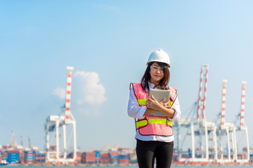 Asian women engineer holding laptop and working with container Cargo freight ship in shipyard at dusk for Logistic Import Export background, safety control. Engineer Concept