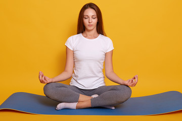 Studio shot of young woman wearing white t shirt and gray leggins, sitting on floor in lotus pose, girl holds hands in praying gesture and keeps eyes closed, posing isolated over yelow background.