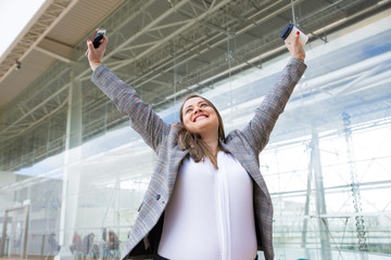 Successful young woman with raised hands posing at office. Smiling businesswoman holding smartphone and paper cup of coffee. Concept of success