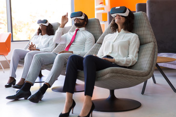Team of excited business colleagues playing VR game. Man and women wearing virtual reality glasses, sitting and relaxing in motion chairs and touching air. VR studio concept
