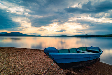 Blue wooden boat moored on the shore of lake under cloudy sky at sunset